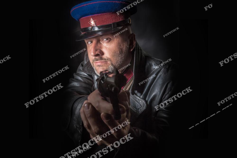 На фотографии изображен офицер советской армии в форме и фуражке, направляющий пистолет в кадр. Его строгий взгляд и кожаная куртка создают образ человека из эпохи СССР. Темный фон и контрастное освещение усиливают атмосферу напряжения и исторической реконструкции.